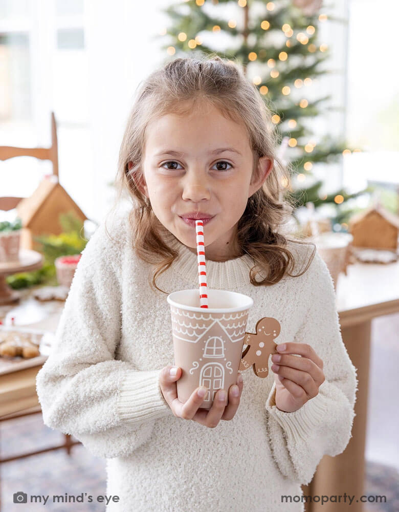 A cute girl in a white sweater holding drinking hot chocolate from Momo Party's 12 oz Gingerbread Handled Paper Cup with a festive gingerbread man handle. She's sipping her drinks with a red and white striped reusable straw. In her back there's a table filled with gingerbread houses and a Christmas tree with lights in background, which creates a cozy yet festive vibe for a whimsical Christmas party for kids this Holiday season.