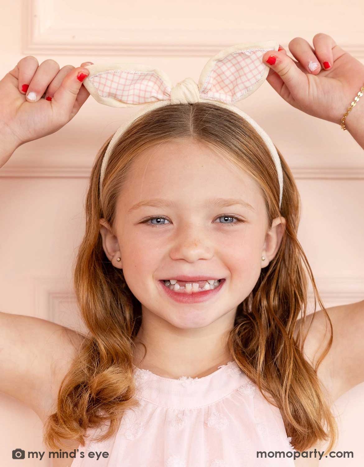 A young girl wearing Momo Party's Gingham Easter Bunny Ear Headband with checkered bow and ears by My Mind's Eye, smiling against a light pink background.