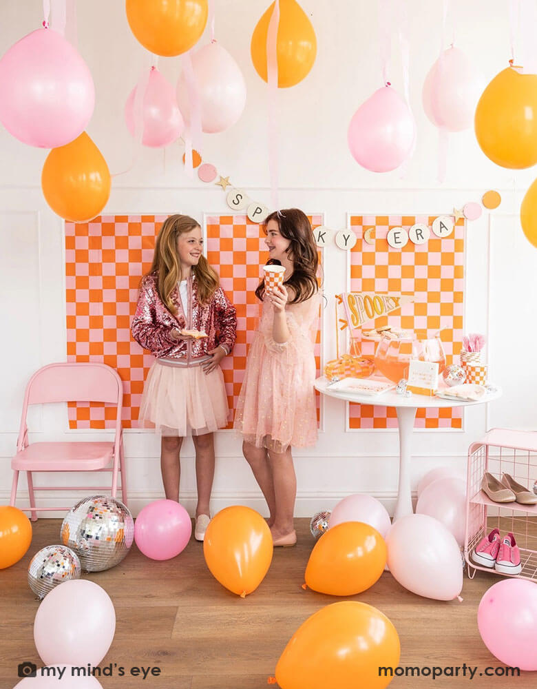 Two middle school girls in their pink outfits chatting with each other in a groovy themed Halloween party decorated with party supplies and decorations in orange and pink. The wall is decorated with orange and pink checkered patterned posters and a Taylor Swift friendship bracelet inspired banner says "Spooky Era." The room is filled with balloons in orange and pink and there's a table full of Halloween party supplies including trendy plates, cups, napkins, pennants from Momo Party.