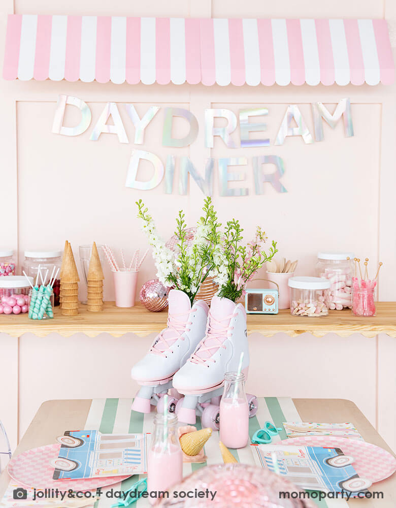 An adorable kid's ice cream themed birthday party tablescape featuring Momo Party's ice cream truck shaped plates, the pink checkered plates, the ice cream icon napkins on a mint striped table runner. On the table there's a roller skate and pink disc balls as centerpiece. Behind the table there's a sundae station with sprinkle toppings, ice cream cones, adorned with a retro pink striped awning as a small ice cream shop, making this a cute and festive ice cream party set up for kids birthday celebration.