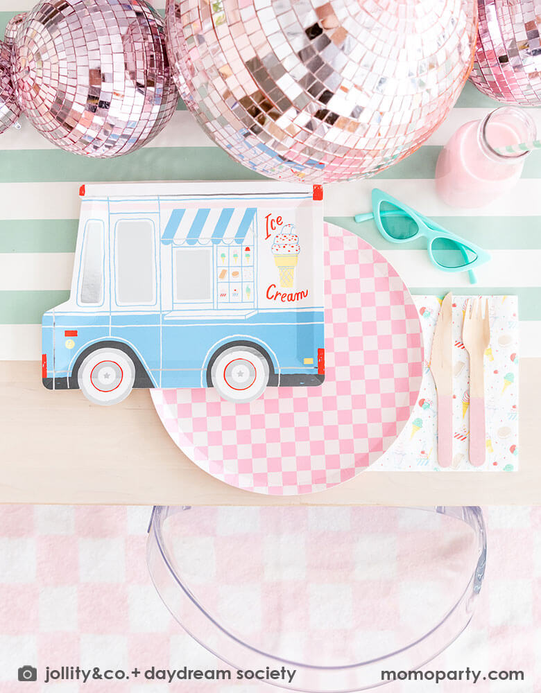An adorable kid's ice cream themed birthday party tablescape featuring Momo Party's ice cream truck shaped plate, the pink checkered plate, the ice cream icon napkin and pink wooden cutlery on a mint striped table runner. Around the placeset there are some summer inspired decoration like a pair of mint sunglasses, pink disco balls, a pair of pink roller skates, some milk bottles with gum ball pink drinks, and some ice cream cone decorations, making this a cute, fun and festive ice cream social.