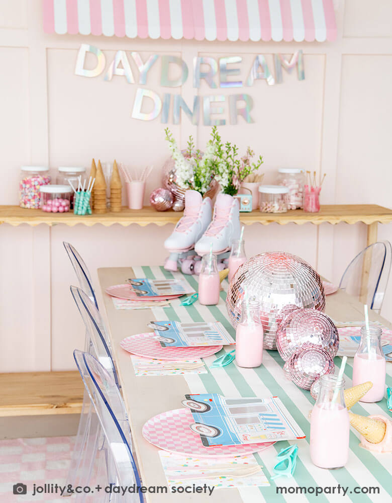 An adorable kid's ice cream themed birthday party tablescape featuring Momo Party's ice cream truck shaped plates, the pink checkered plates, the ice cream icon napkins on a mint striped table runner. On the table there's a roller skate and pink disc balls as centerpiece. Behind the table there's a sundae station with sprinkle toppings, ice cream cones, adorned with a retro pink striped awning as a small ice cream shop, making this a cute and festive ice cream party set up for kids birthday celebration.