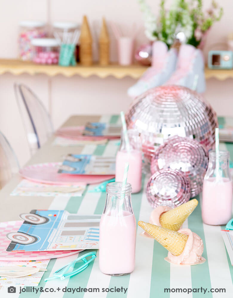 An adorable kid's ice cream themed birthday party tablescape featuring Momo Party's ice cream truck shaped plates, the pink checkered plates, the ice cream icon napkins on a mint striped table runner. On the table there's a roller skate, pink disc balls and ice cream cone decorations as the centerpiece. Behind the table there's a sundae station with sprinkle toppings, ice cream cones, making this a cute and festive ice cream party set up for kids birthday celebration.