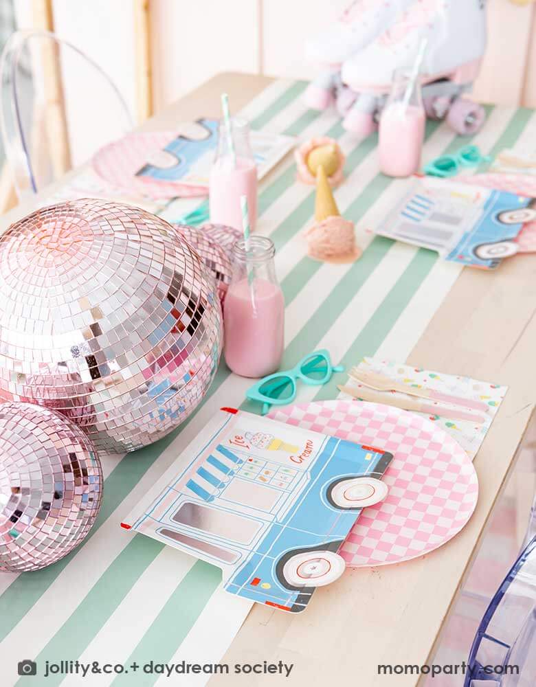 An adorable kid's ice cream themed birthday party tablescape featuring Momo Party's ice cream truck shaped plate, the pink checkered plate, the ice cream icon napkin and pink wooden cutlery on a mint striped table runner. Around the placeset there are some summer inspired decoration like a pair of mint sunglasses, pink disco balls, a pair of pink roller skates, some milk bottles with gum ball pink drinks, and some ice cream cone decorations, making this a cute, fun and festive ice cream social.