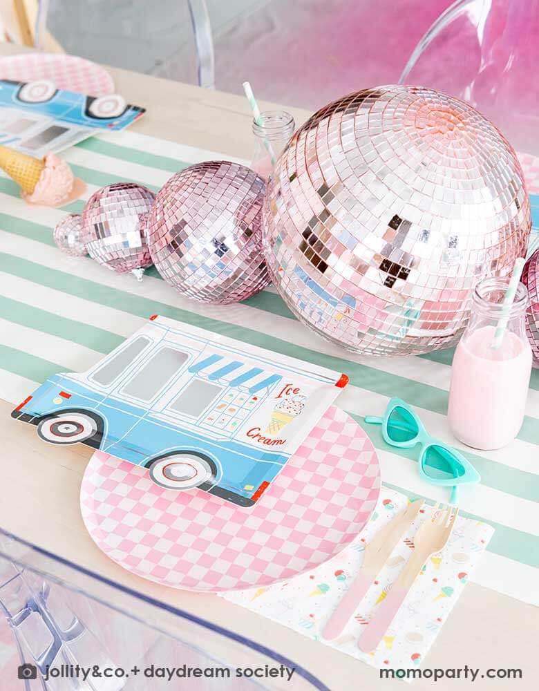 An adorable kid's ice cream themed birthday party tablescape featuring Momo Party's ice cream truck shaped plate, the pink checkered plate, the ice cream icon napkin and pink wooden cutlery on a mint striped table runner. Around the placeset there are some summer inspired decoration like a pair of mint sunglasses, pink disco balls, a pair of pink roller skates, some milk bottles with gum ball pink drinks, and some ice cream cone decorations, making this a cute, fun and festive ice cream social.