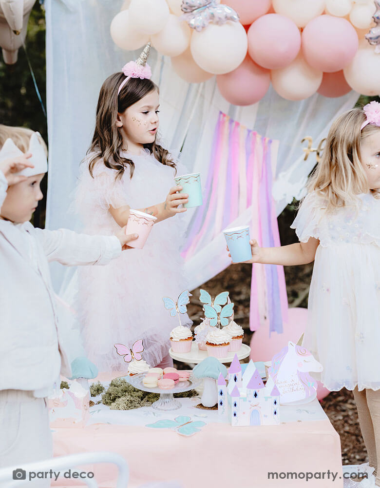 A few kids dressed in beautiful dresses in a dreamy fairytale themed birthday party table featuring Momo Party's unicorn princess fairy themed party including butterfly cupcake toppers, castle centerpiece and butterfly shaped napkins. The kids are holding Momo Party's 7.4 oz pastel bow paper cups in 3 different colors. In the back there's a pastel balloon cloud adorned with butterfly foil balloons and a white curtain, making this a whimsical set up for kid's fairytale birthday celebration.