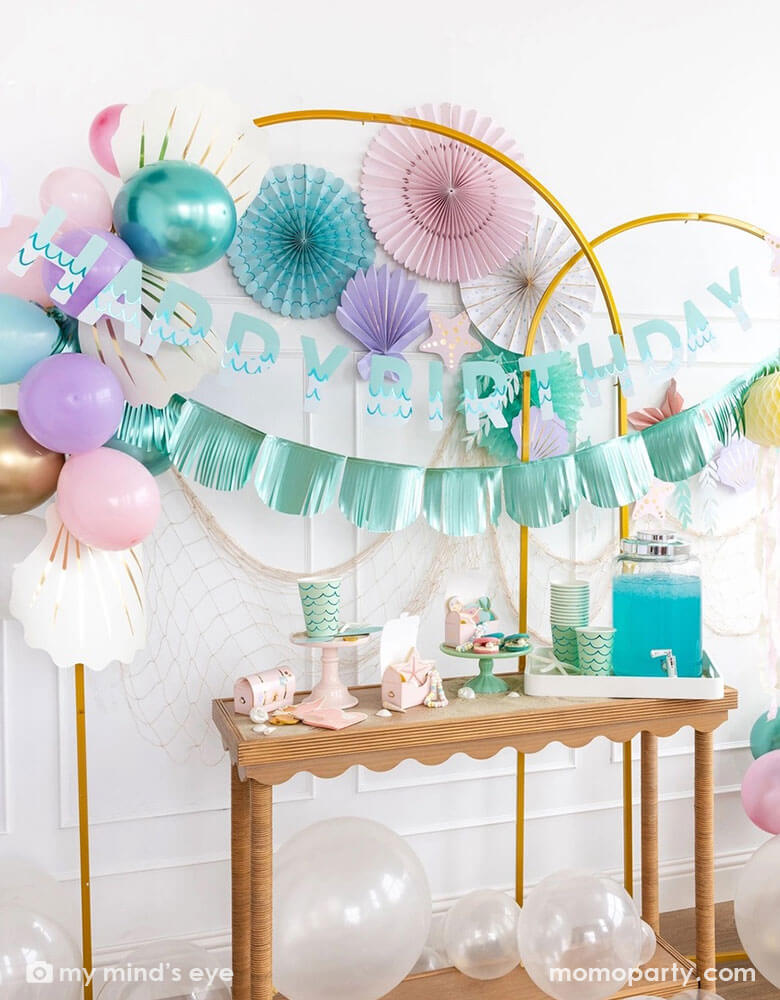 An enchanting mermaid party set up features Momo Party's mermaid decorations and and supplies by My Mind's Eye including including the Under the Sea paper fan set in pastel colors of light pink, aqua, teal, lilac and cream, Mermaid Happy Birthday Banner Set with aqua foil fringe banner and shell shaped paper decorations on the wall. On the table there are various mermaid themed tableware including cups, napkins, plates and treat boxes. On the floor are clear latex balloons creating a whimsy bubble scene.