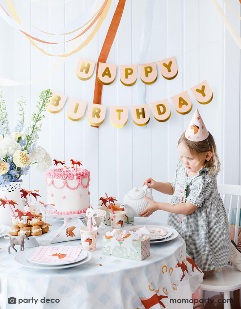 A preschool girl with a birthday party hat in a gingham dress pouring tea from a tea pot on a party table featuring Momo Party's floral pony themed party supplies including floral pony party cups, plates, cupcake toppers, napkins treat boxes and paper straws. In the middle of the table is a pink and white buttercream cake topped with pony shaped birthday candles, along with the blush happy birthday party banner hung on the wall - making this a perfect inspo for a girl's horse themed birthday party.