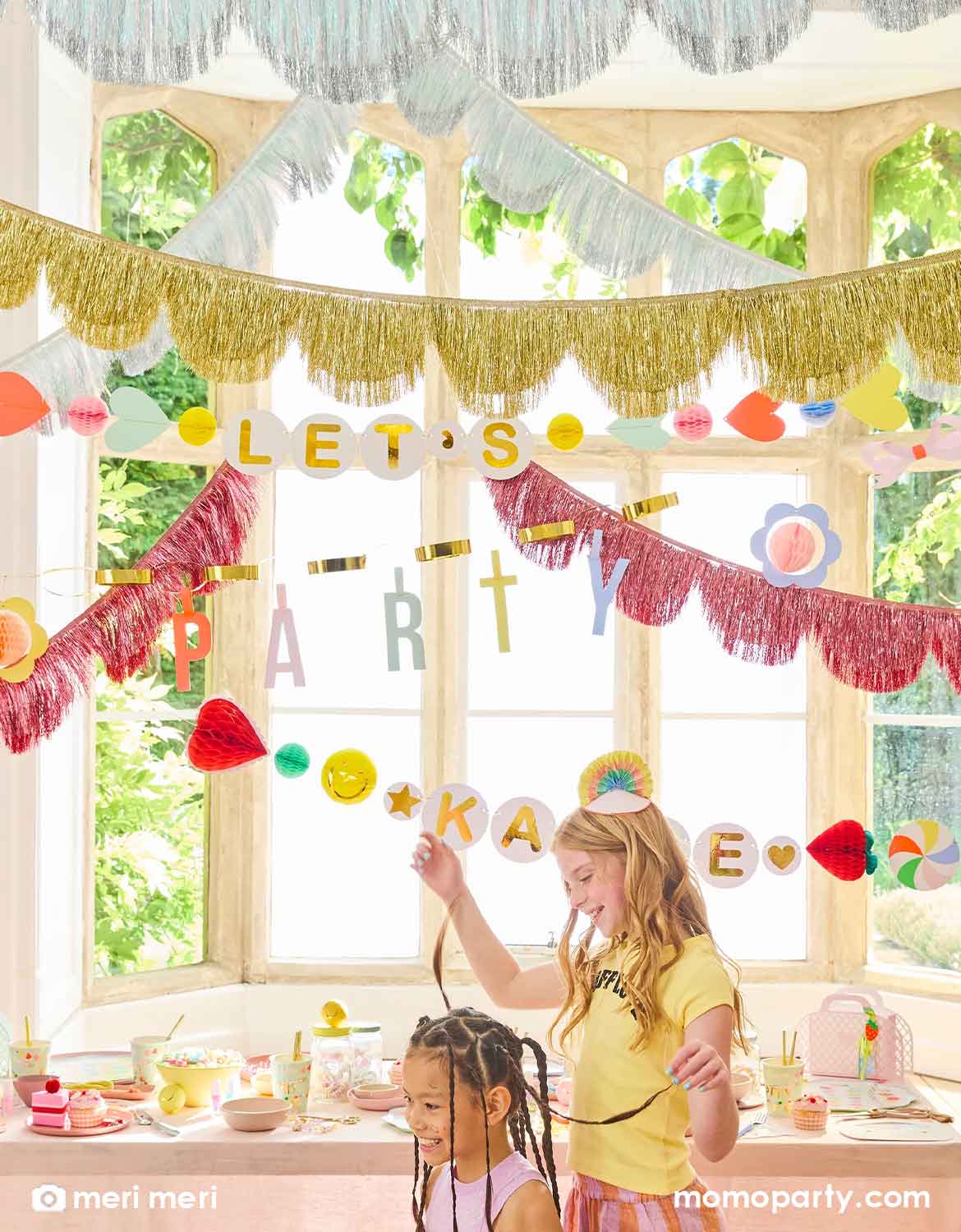 Two pre-teen girls in a room decorated with colorful Momo Party's Charm Party Collection by Meri Meri including festive banners, metallic fringe garlands, and a table of charm party themed tablewares including plates, napkins and cups. A perfect set up for girl's sleepover party or a BFF gathering.
