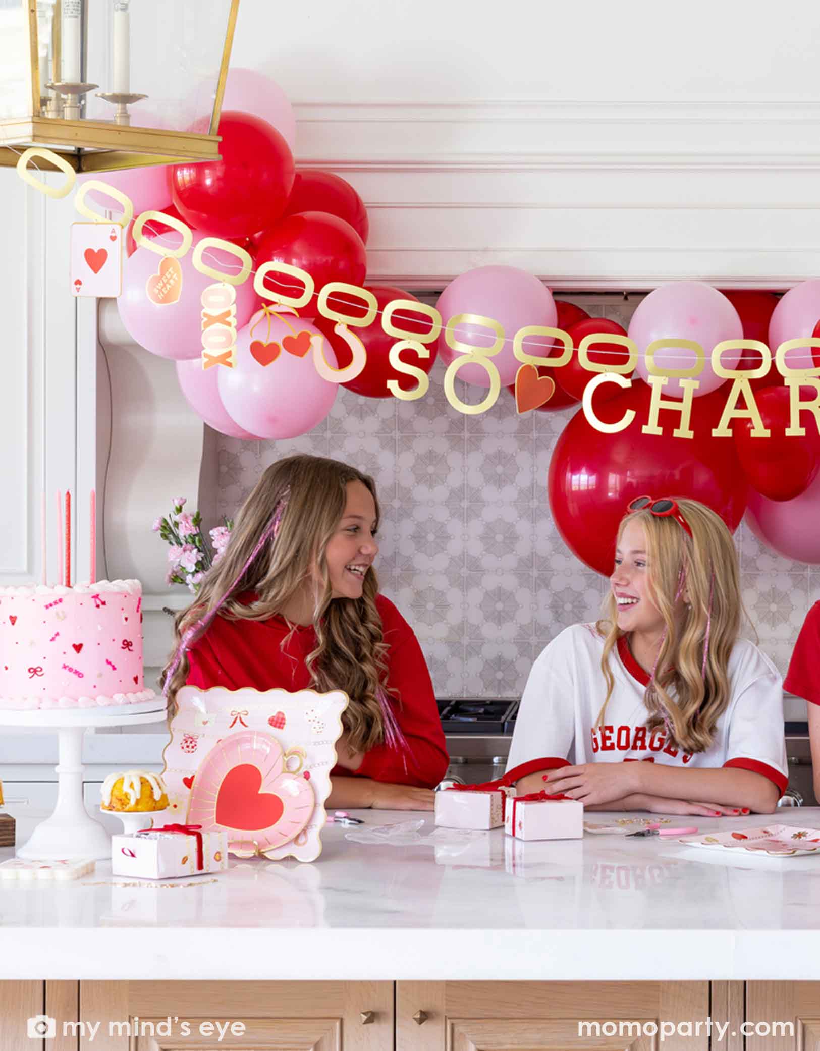 Two girls sitting at a Valentine's Day party table filled with heart themed party supplies from Momo Party including plates, napkins, and a cake. In the back there's a heart-themed balloon arch in red and pink and a gold foil party banner spells "So Charming".
