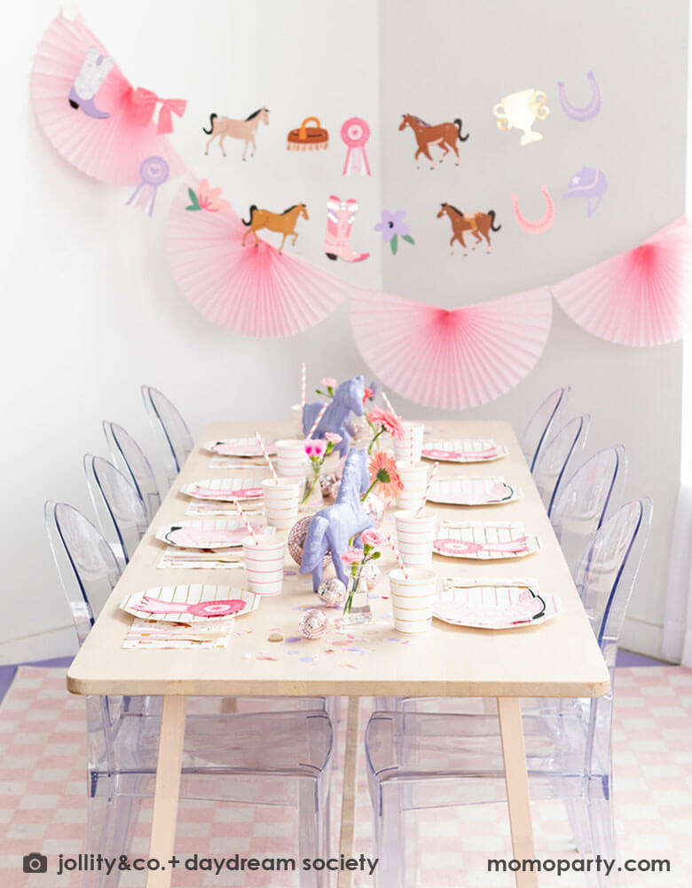 A girl's rodeo themed party decoration set up features Momo Party's cowgirl pony themed party supplies and decorations by Daydream Society including cowgirl boots shaped napkins, show ribbon shaped napkins, cowgirl rodeo themed paper napkins and pink striped paper plates and cups. With cowgirl themed party garland featuring paper pennants including ponies, show ribbons, pink horseshoes, bowes and flowers hung above the table, makes this a perfect inspiration for a girly rodeo themed celebration!