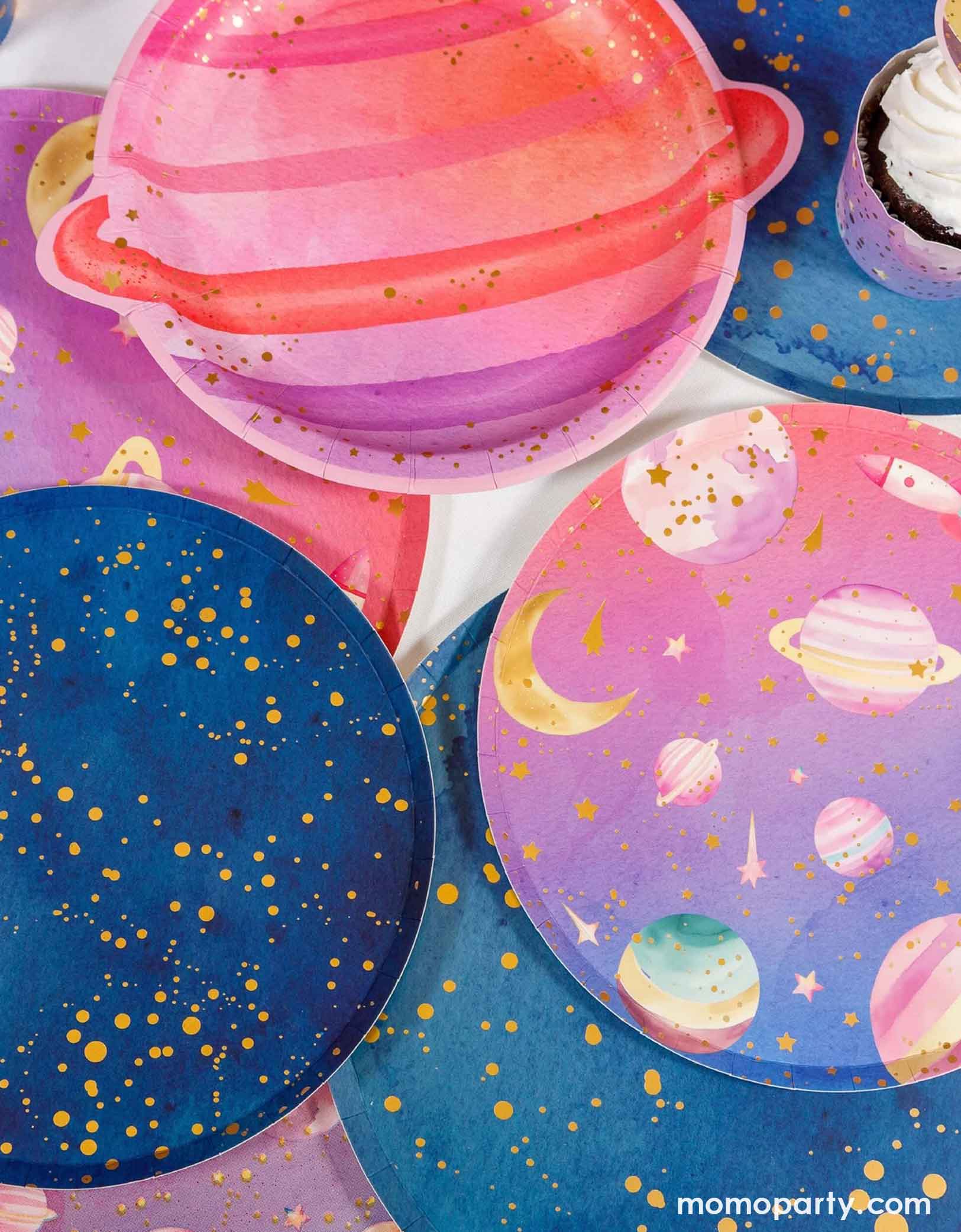 Various space themed party plates from Momo Party on a table including a pink planet shaped plate, blue galaxy plates with gold dots on them, and pink and lilac ombre space plates.