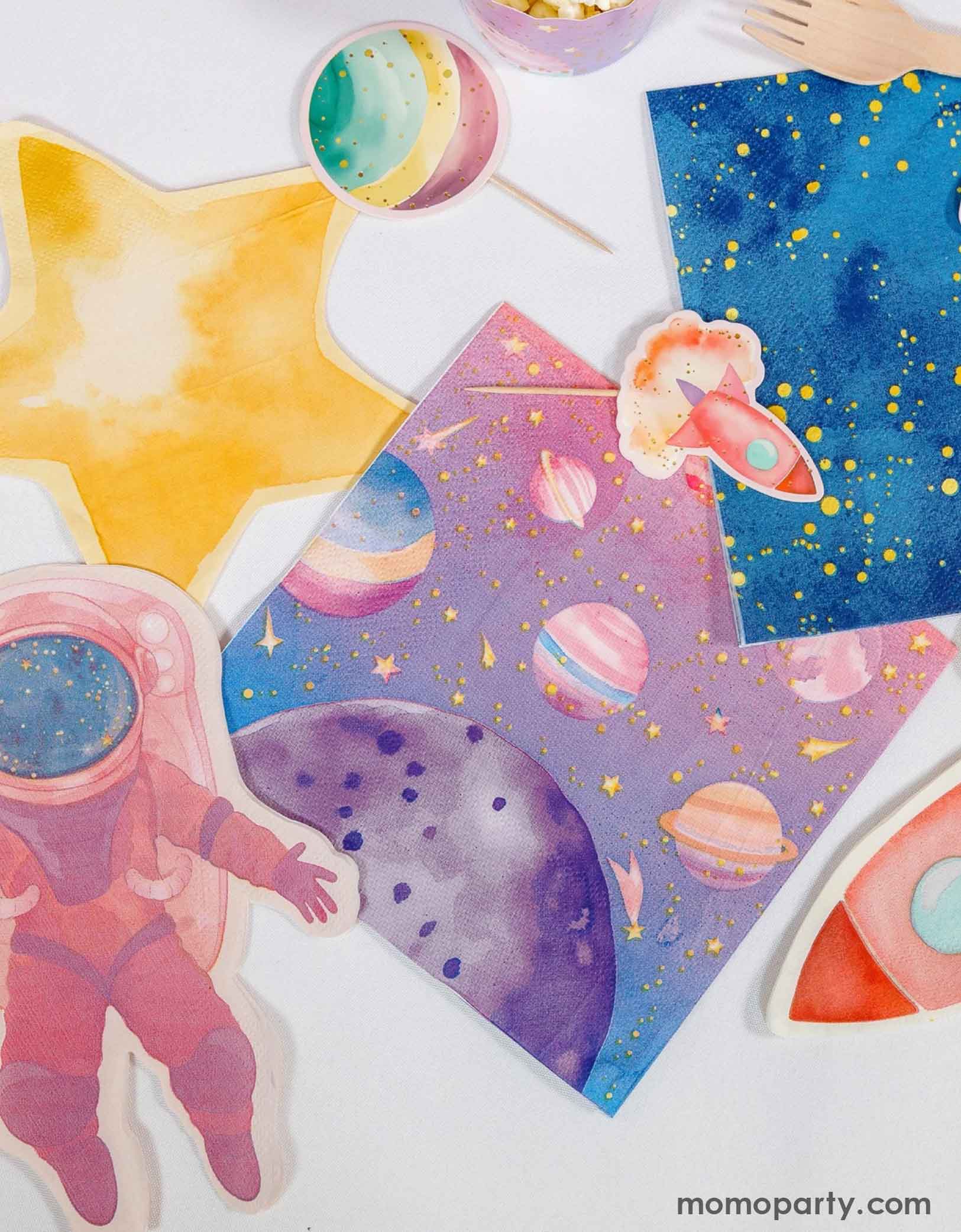Colorful space-themed party napkins including stars, planets, rocket ship and an astronaut shaped napkin from Momo Party spread out along with some space themed cupcake toppers on a white background.
