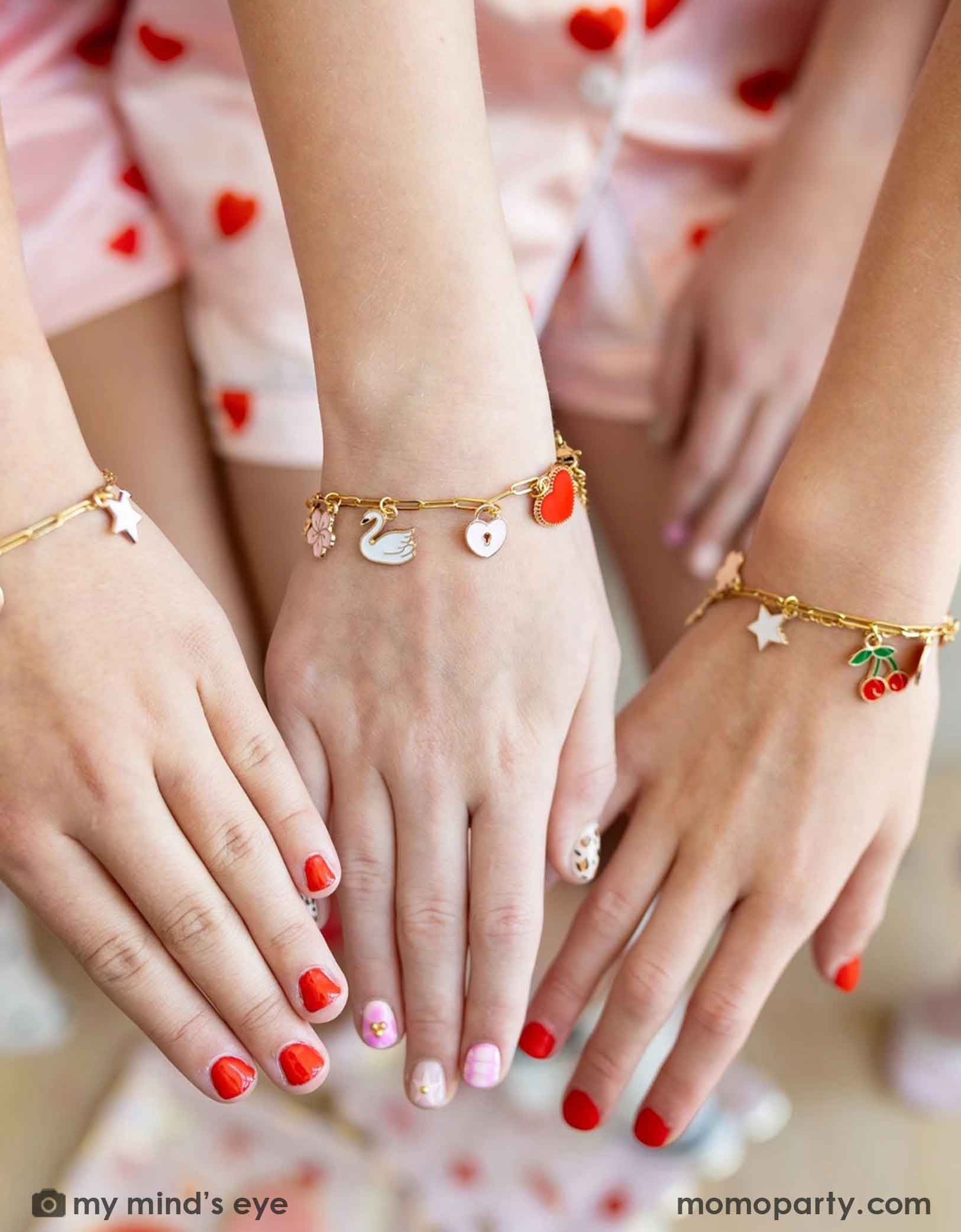 Three girls' hands with red nail polish showing their gold bracelets with various charms from Momo Party against a blurred background at a Valentine's Day themed sleep over party celebration.