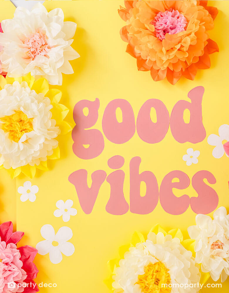 A yellow party backdrop wall with with GOOD VIBES in retro font adorn with cream, pink and orange tissue paper pom pom flower decorations from Momo Party.