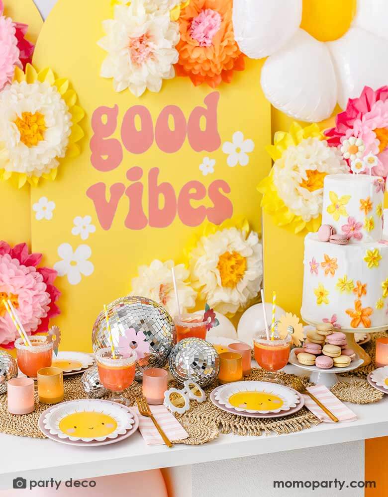 A "Good Vibes" themed party featuring Momo Party's scallop edged sun plates, pink and white striped napkins and sunflower shaped sunglasses. On the table is a two-tier butter cream birthday cake decorated with bright flowers in orange, yellow and peach colors, along with some color-matching macarons and drinks and fun disco balls as the centerpiece. In the back is a yellow backdrop with GOOD VIBES in retro font adorn with tissue paper pom pom flower decorations.