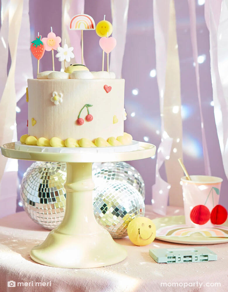 A festive retro themed birthday party table featuring a pale buttercream birthday cake topped with Momo Party's Happy Face Icons candle which includes a strawberry, a rainbow, a smiley face, a heart, and a couple of flowers. On the table there are a few disco balls and cassette tapes as the centerpiece, along with Meri Meri's happy faces cheery honeycomb paper cup and a rainbow napkin, with pastel streamers in the back, makes this a great inspo for a fun 90s' retro themed party.