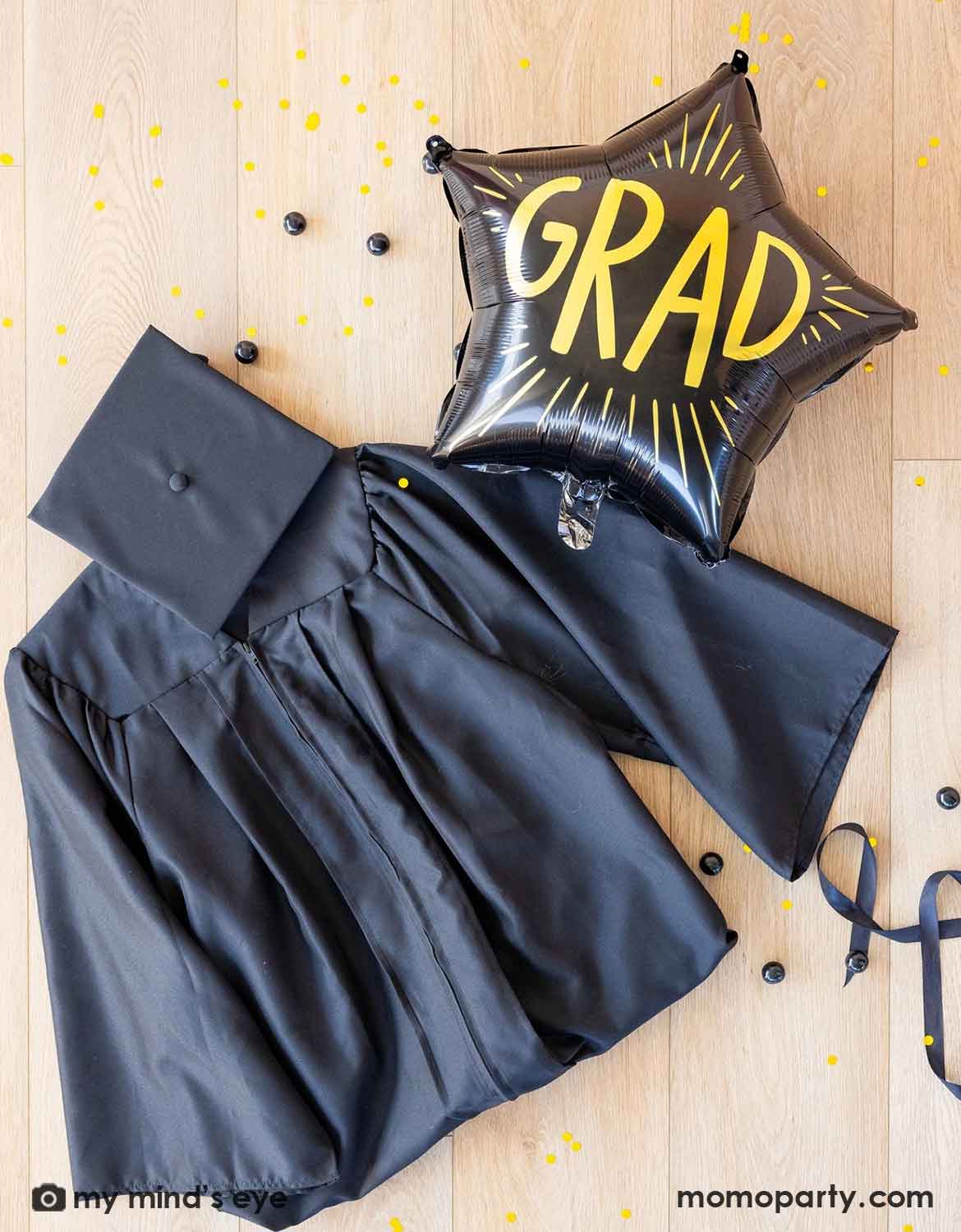 An overshot of a graduation gown, cap, and Momo Party's 24" black star-shaped 'GRAD' foil balloon on a wooden floor with some black ribbons and black gum balls around.