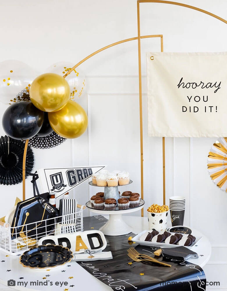 A graduation party featuring Momo Party's graduation party supplies by My Mind's Eye including U did it Grad felt party pennant, class of 2024 black striped dinner napkins, graduation cap shaped plates. On the table there are some cupcakes, cookies and popcorn on a black congratulation paper table runner and some black grad party cups and grad cap shaped napkins. In the back there are a gold backdrop frame adorned with some black and gold balloons and party fans, along with a "Hooray You Did It!" banner.