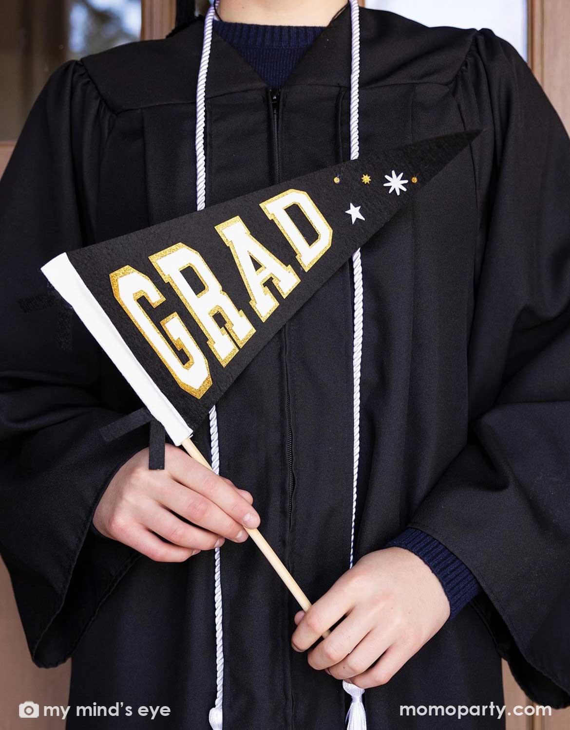 A collage grad in his graduation gown and cap holding Momo Party's black Grad Felt Pennant Banner with 'GRAD' tex on it standing by a black door at school.