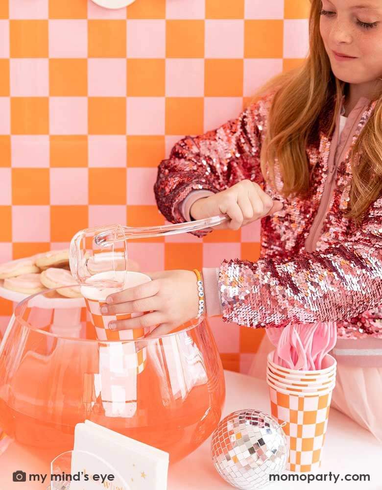 A girl in her pink sequin jacket stands in front of a wall covered with an orange and pink wallpaper. She's pouring some punch into Momo Party's 10oz orange and pink checkered party cup, next to the punch bowl is a disco ball decoration and more checkered cups with pink utensils for a groovy themed Halloween party in this spooky season.