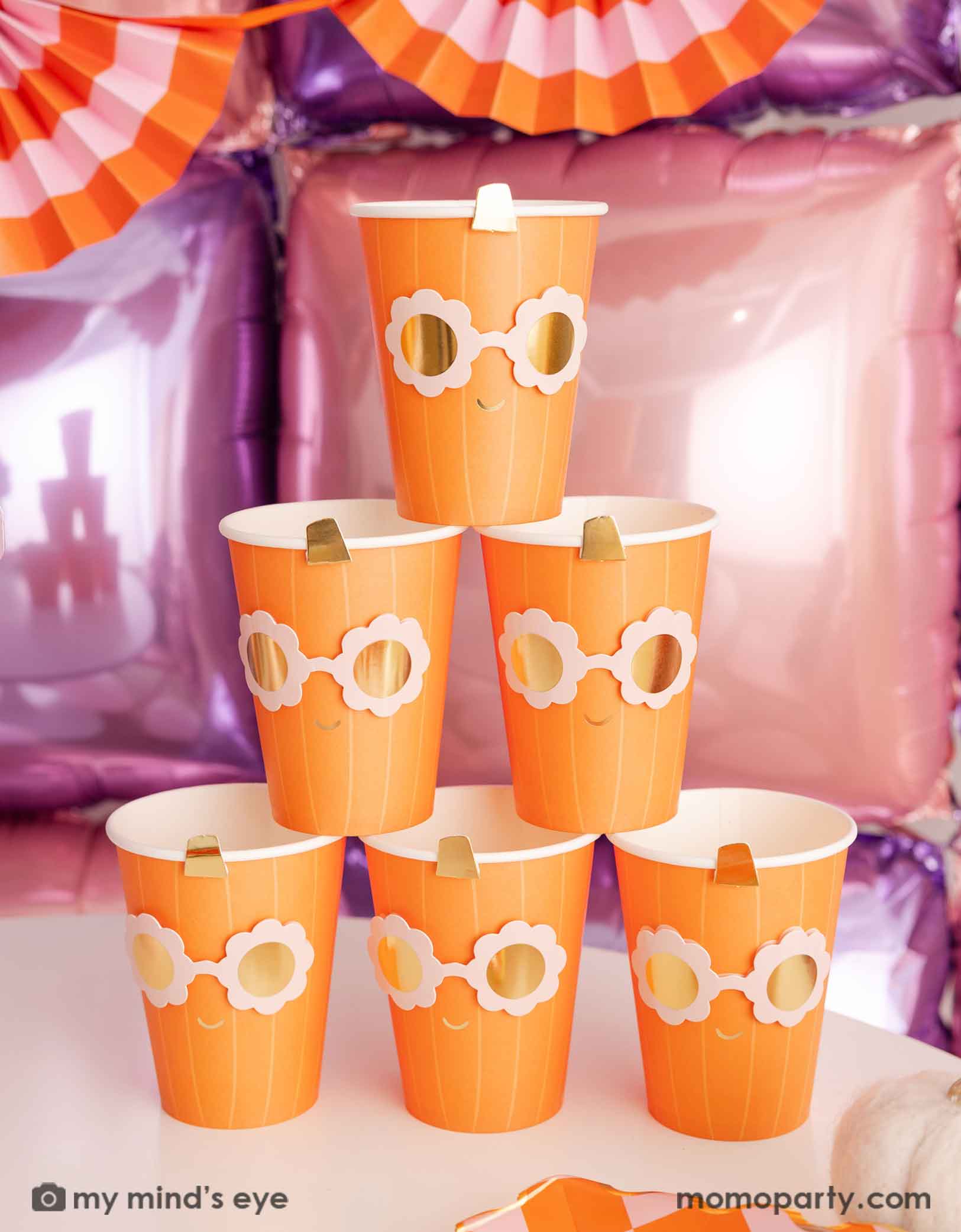 A stack of the orange pumpkin shaped paper party cups with daisy-shaped sunglasses from Momo Party on a Halloween inspired background with a foil balloon wall in pink and lilac and an orange and pink bunting garland.