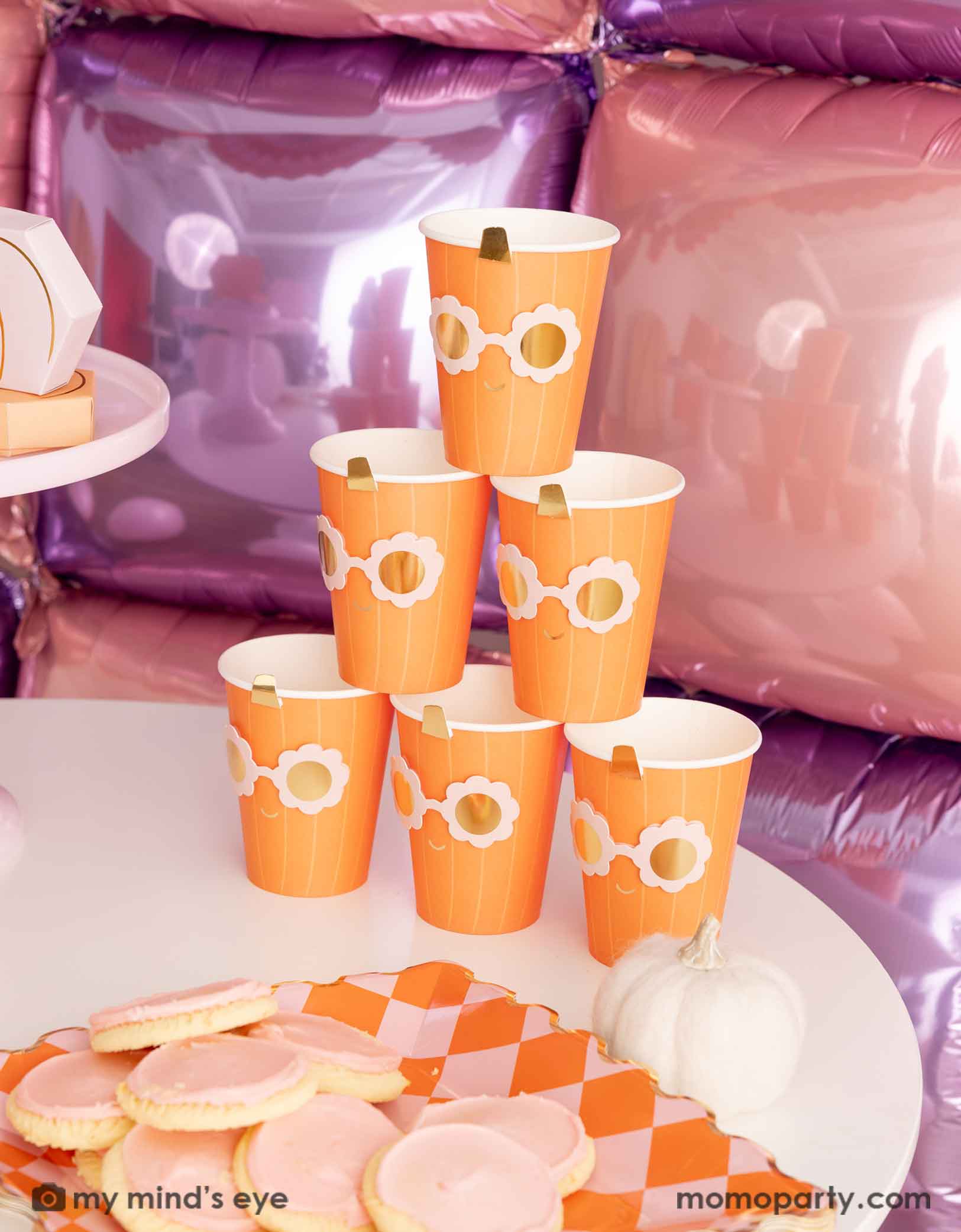A stack of the orange pumpkin shaped paper party cups with daisy-shaped sunglasses from Momo Party on a Halloween inspired background with a foil balloon wall in pink and lilac. On the table there's a orange and pink checkered dinner plate filled with pink sugar coated cookies for a party.