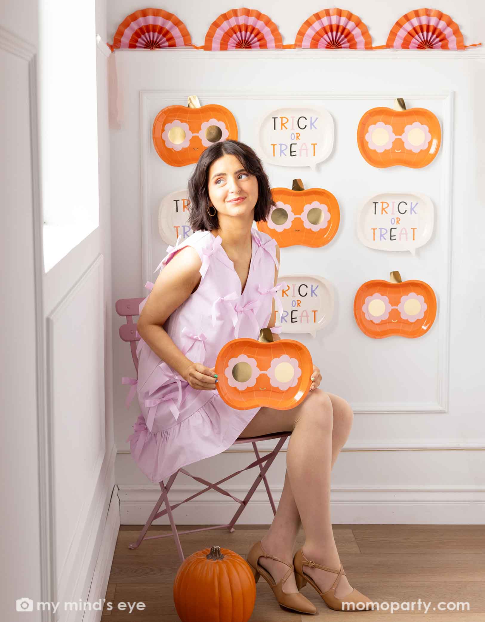 A woman in her pink dress sitting on a chair holding Momo Party pumpkin-shaped plate with daisy shaped sunglasses, with a white wall decorated with Momo Party's groovy halloween pumpkin shaped plates and trick or treat plates in the background.