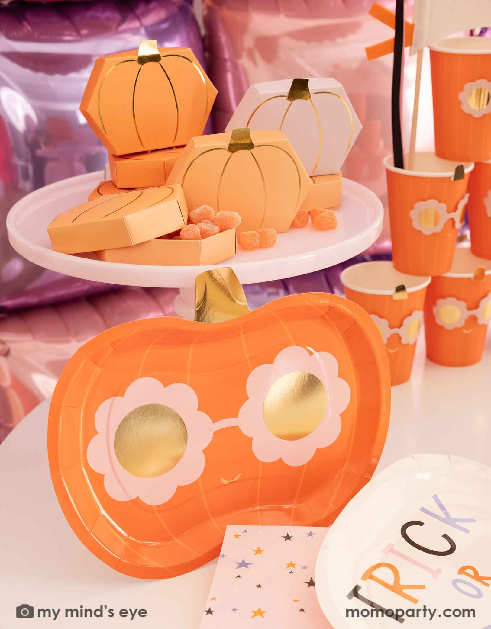 A Halloween party table featuring various Halloween-themed party supplies from Momo Party including pumpkin-shaped plates, party cups, trick or treat plates, napkins and some pumpkin shaped treat boxes filled with orange gummies on a pink cake stand, in front of a pink and lilac balloon wall background.