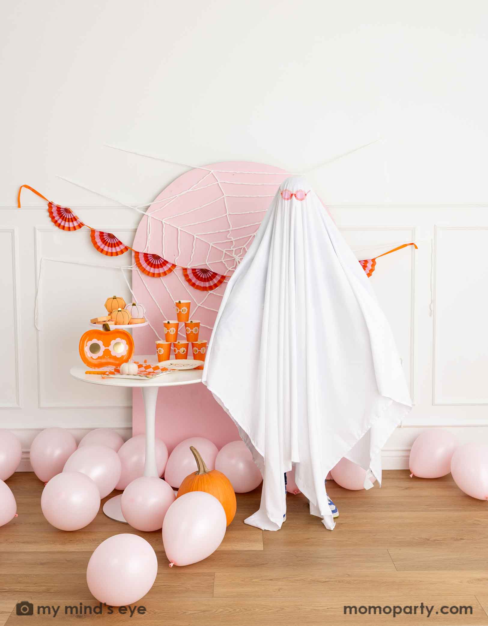 A kid's friendly Halloween party setup featuring a pale pink wooden curved backdrop board decorated with spider web, a party table filled with groovy Halloween pumpkin party tablewares from Momo Party and a bunch of pink balloons on the floor. A kid dresses as a sheet ghost wearing a pink sunglasses, making it groovy and fun vibe for this Halloween celebration.