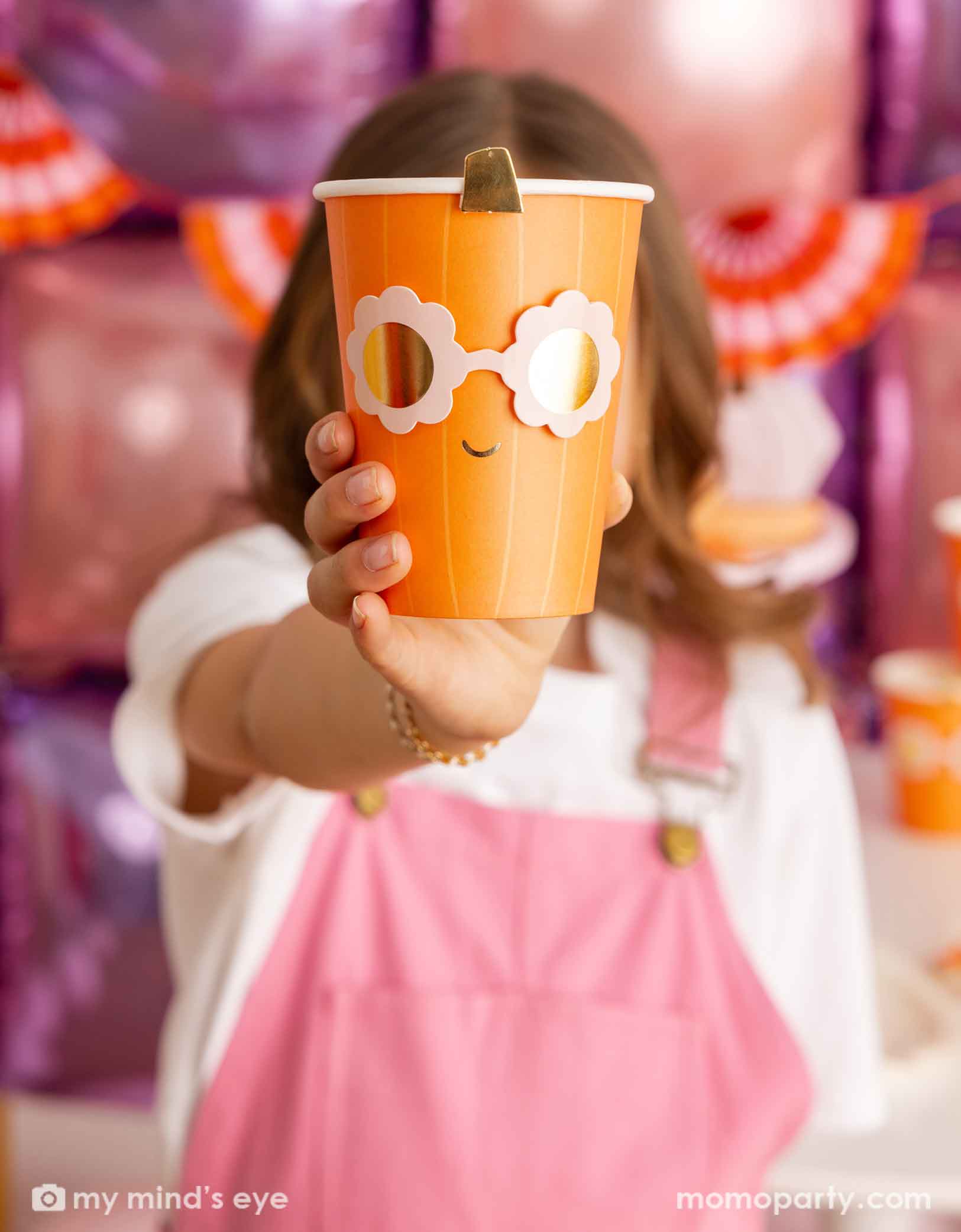 A girl in a pink overalls holding an orange pumpkin shaped paper party cup with a pair of daisy-shaped sunglasses from Momo Party in a festive Halloween setting featuring a balloon wall and an orange and pink bunting banner.