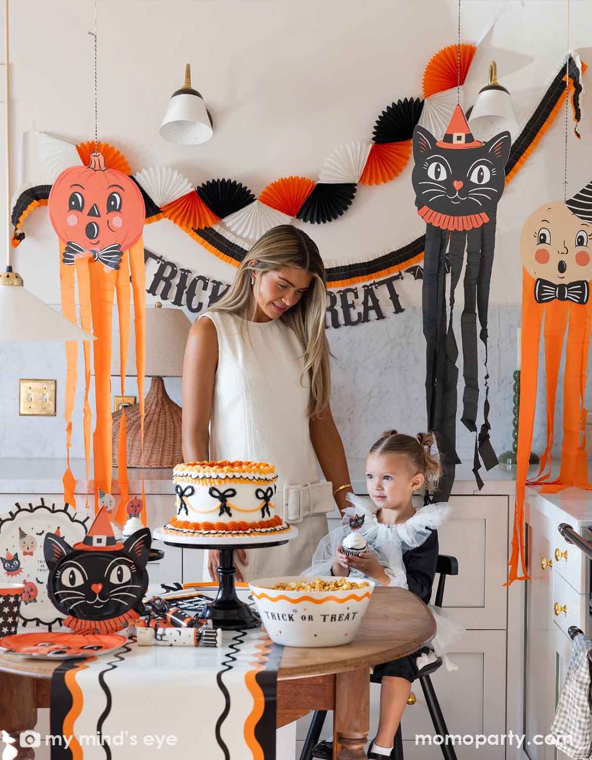 A woman and a young girl celebrate Halloween in a decorated kitchen featuring various vintage Halloween themed party supplies and decorations from Momo Party including huge hanging decorations, trick or treat banner, bunting garlands, and a table with a Halloween-themed cake, a bowl of candy corn, and vintage Halloween themed party plates, serving bowls, cups, napkins, cupcake toppers and a table runner, making this a fun and festive set up for kids and family in the spooky season.