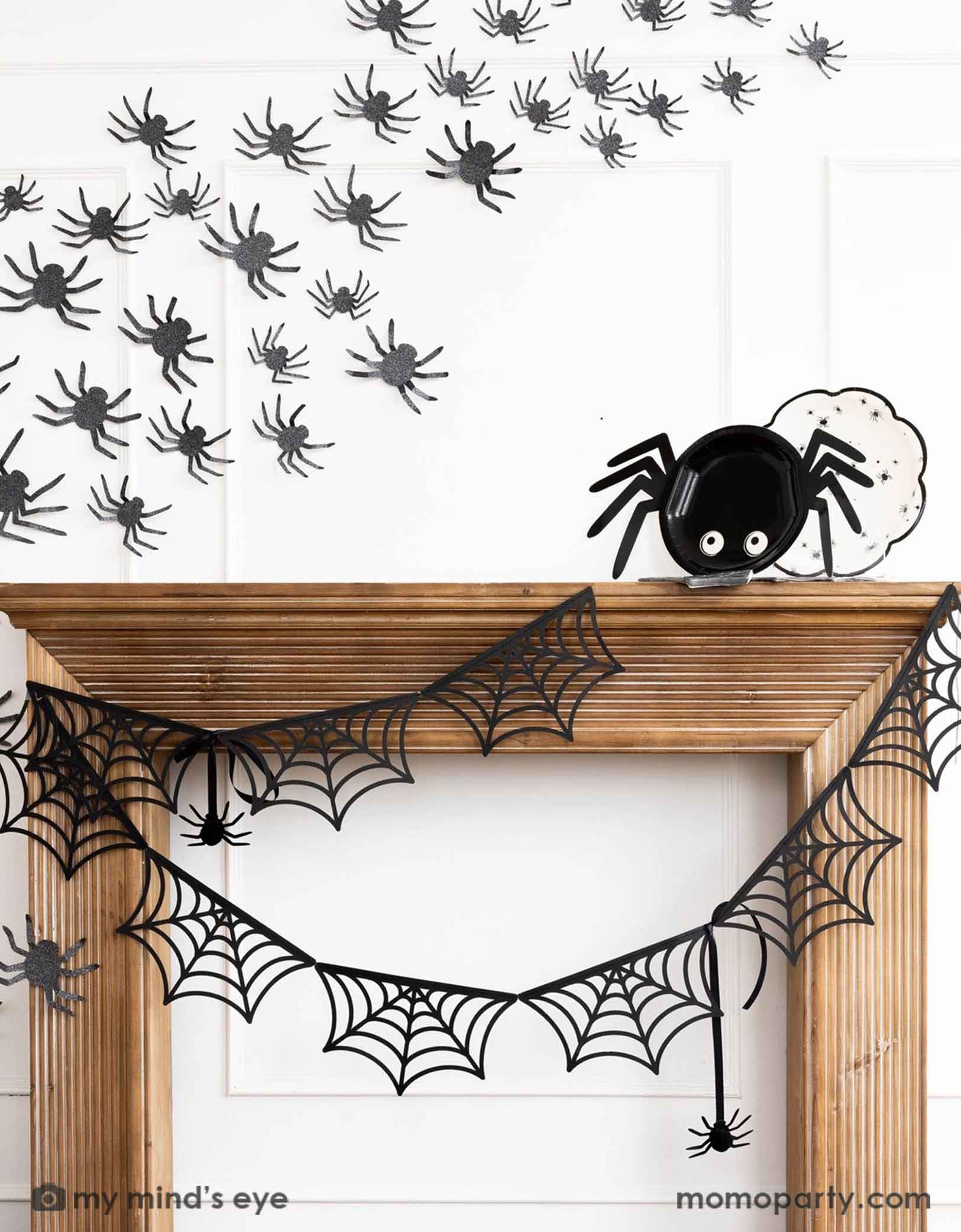 A wooden mantel against a white wall decorated with Halloween decorations from Momo Party including the spider web banner, black spider decorations on the wall and the 14" x 7" spider shaped plate and a round plate with spider designs on it. Making this a kid's friendly cute Halloween decorating idea for the spooky season.