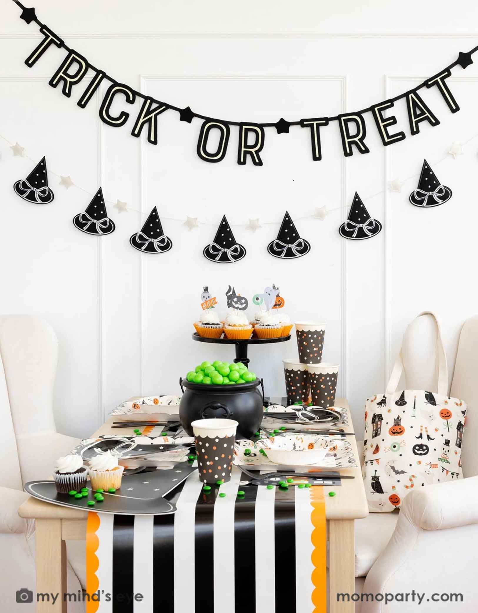 A Halloween-themed party with Momo Party's 6 ft 'Trick or Treat' glow in the dark banner and the witch's hat garland hung on the white wall. There's a table decorated with multiple Halloween party supplies by Momo Party including witch's hat shaped bamboo tray, party cups and orange, black and white striped table runner draping from the table which some orange and black balloons next to. A cute and simple set up for kids to celebration Halloween at home.