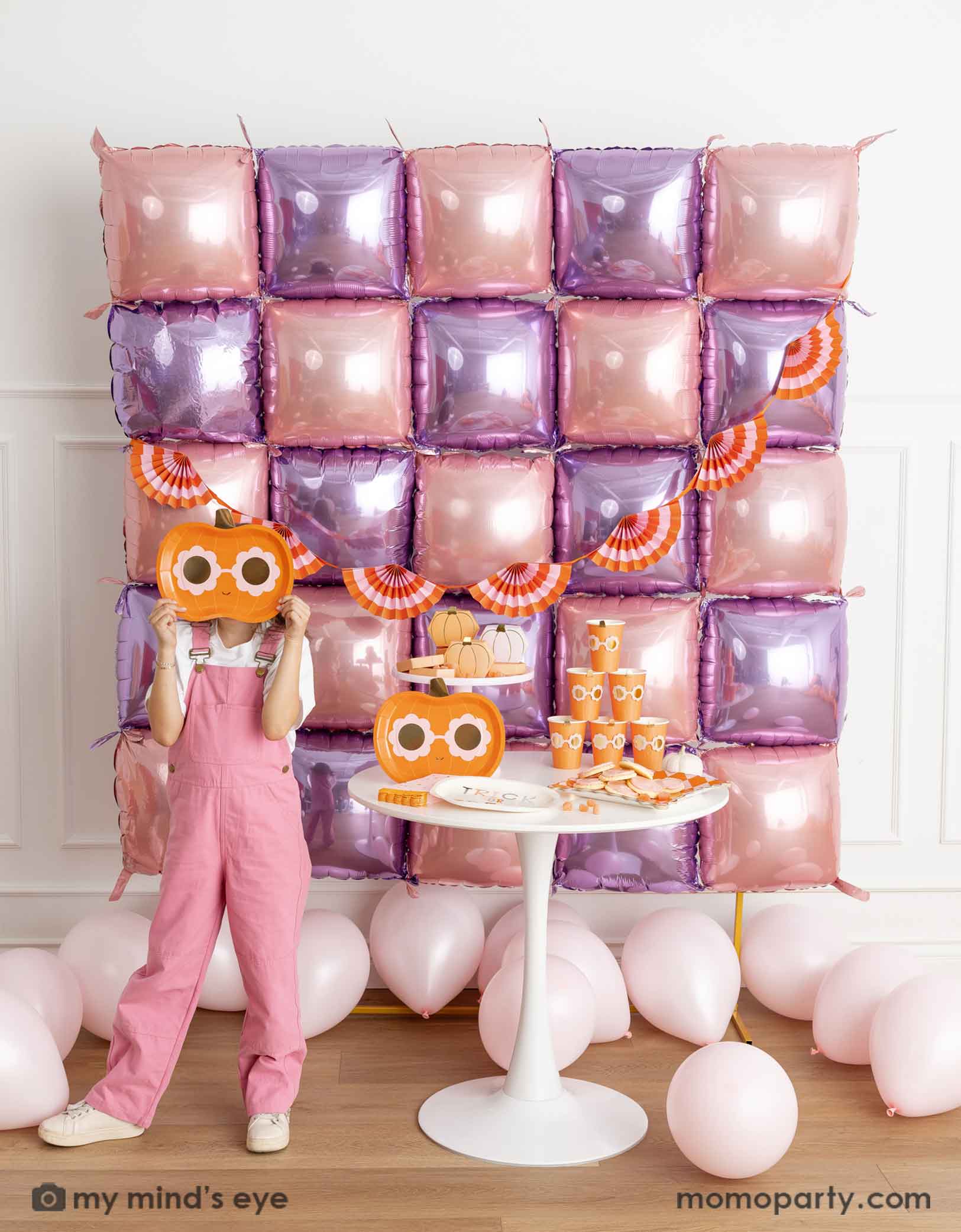 A girl in her pink overalls holding Momo Party's pumpkin-shaped plate standing in front of a decorative wall with pink and purple foil balloons adorned with an orange and pink bunting garland. Next to her is a white round table displaying groovy halloween pumpkin party tablewares including party plates, cups, napkins and more. On the floor are some pale pink latex balloon, making this a festive kid's friendly pink Halloween celebration.