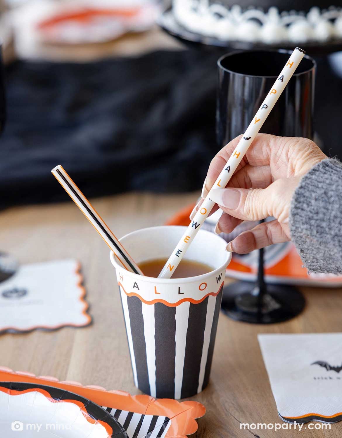 A hand holds a white paper straw printed with small orange and black texts of Happy Halloween next to Momo Party's black and white striped paper cup on a wooden table, with Halloween party supplies including plates and napkins and decorations in the background.
