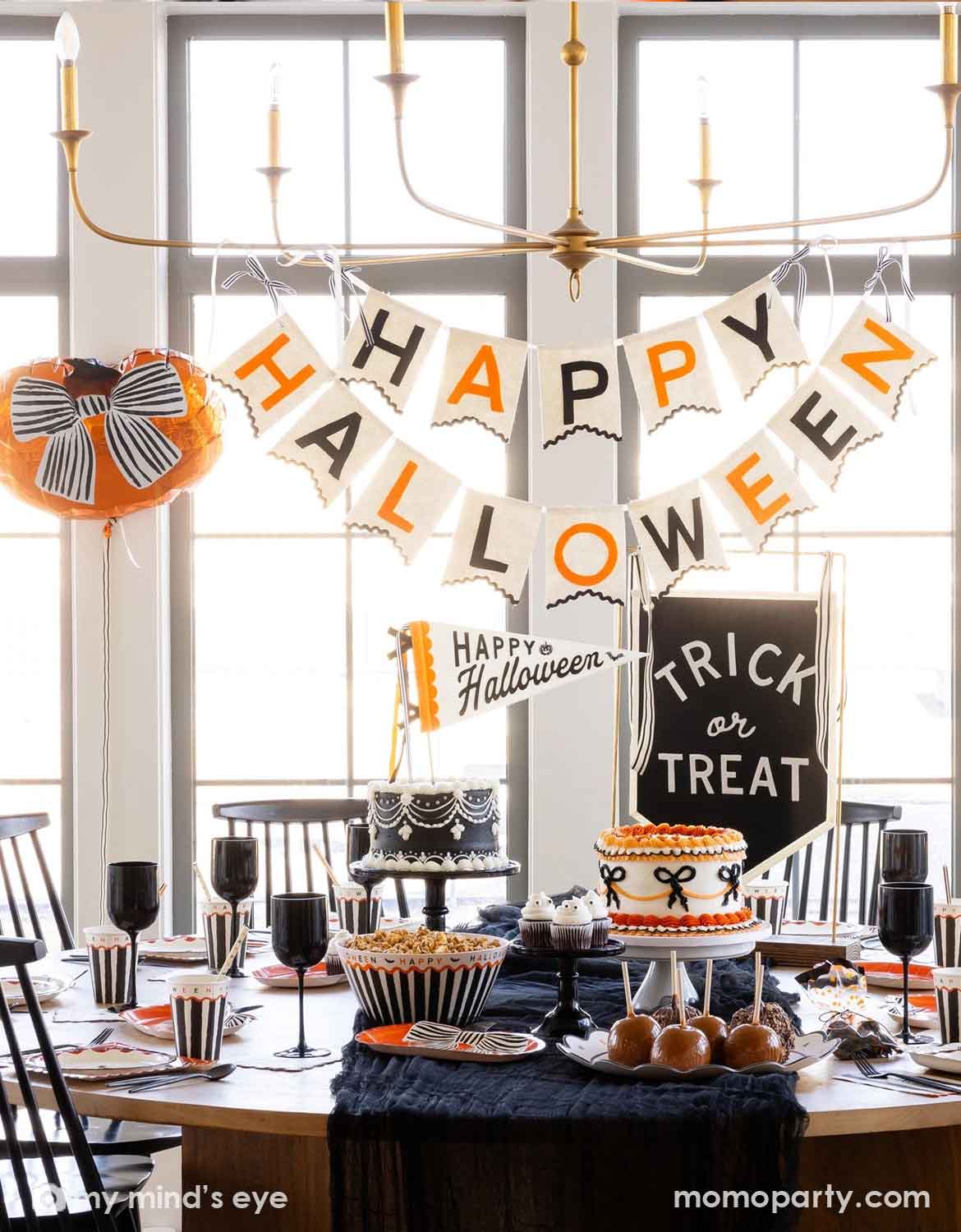 A styled Halloween party table setup in a bright dining room. Momo Party's Halloween large banner reading HAPPY HALLOWEEN hangs above the table, with a smaller sign that says Happy Halloween and a chalkboard sign that says Trick or Treat. On the table various Halloween themed party tablewares are featured, including party cups, plates, napkins and serving bowls - making this a modern and elegant Halloween party decorating inspo.