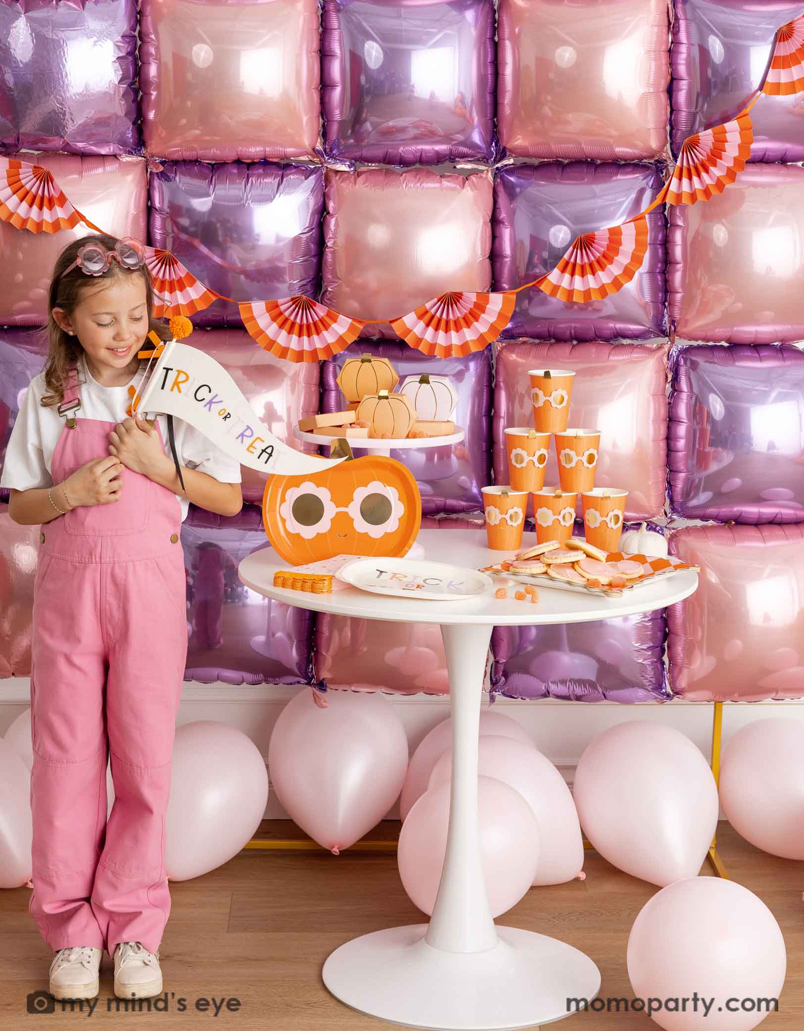 A girl in her pink overalls holding Momo Party's trick or treat felt party pennant standing in front of a decorative wall with pink and purple foil balloons adorned with an orange and pink bunting garland. Next to her is a white round table displaying groovy halloween pumpkin party tablewares including party plates, cups, napkins and treat boxes. On the floor are some pale pink latex balloon, making this a festive kid's friendly pink Halloween celebration.