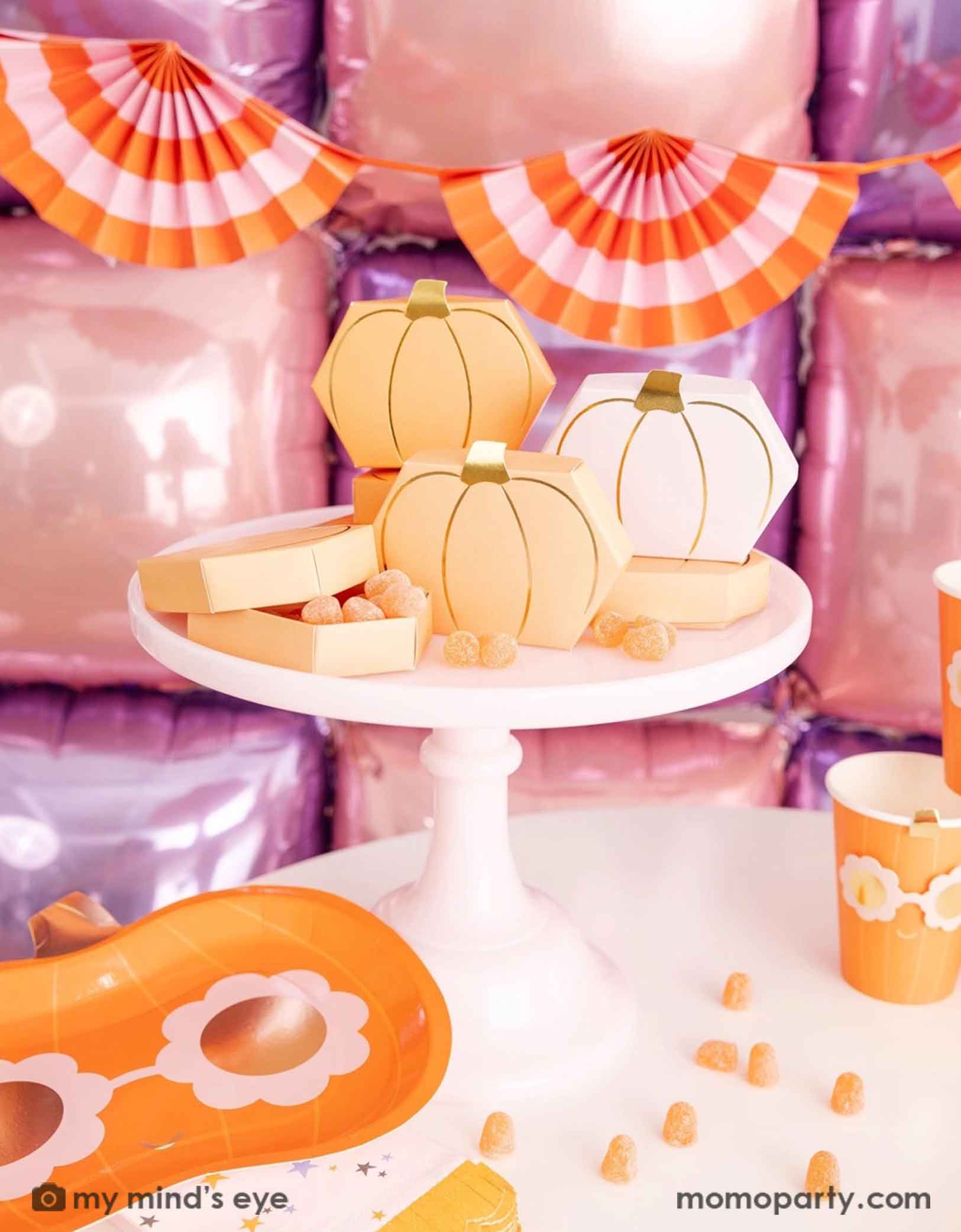 A festive Halloween-themed party setup with Momo Party's Pumpkin-shaped boxes by My Mind's Eye filled with orange gummies on a pale pink cake stand with festive pink Halloween decorative background with foil balloon wall and bunting garland. On the table there are groovy pumpkin shaped plates and party cups and some candy corn spread-out. A sweet and kid's friendly set up for the spooky season.