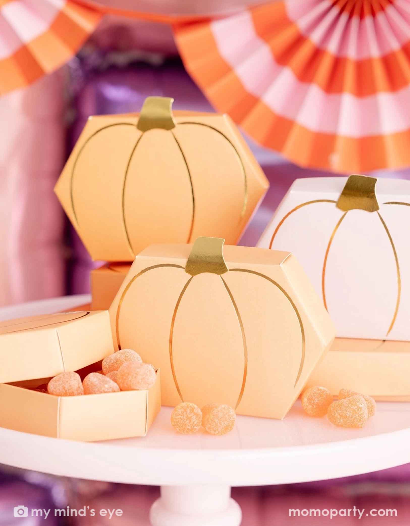 A close up of Momo Party's Pumpkin-shaped boxes by My Mind's Eye filled with orange gummies on a pale pink cake stand with festive pink Halloween decorative background with foil balloon wall and bunting garland.