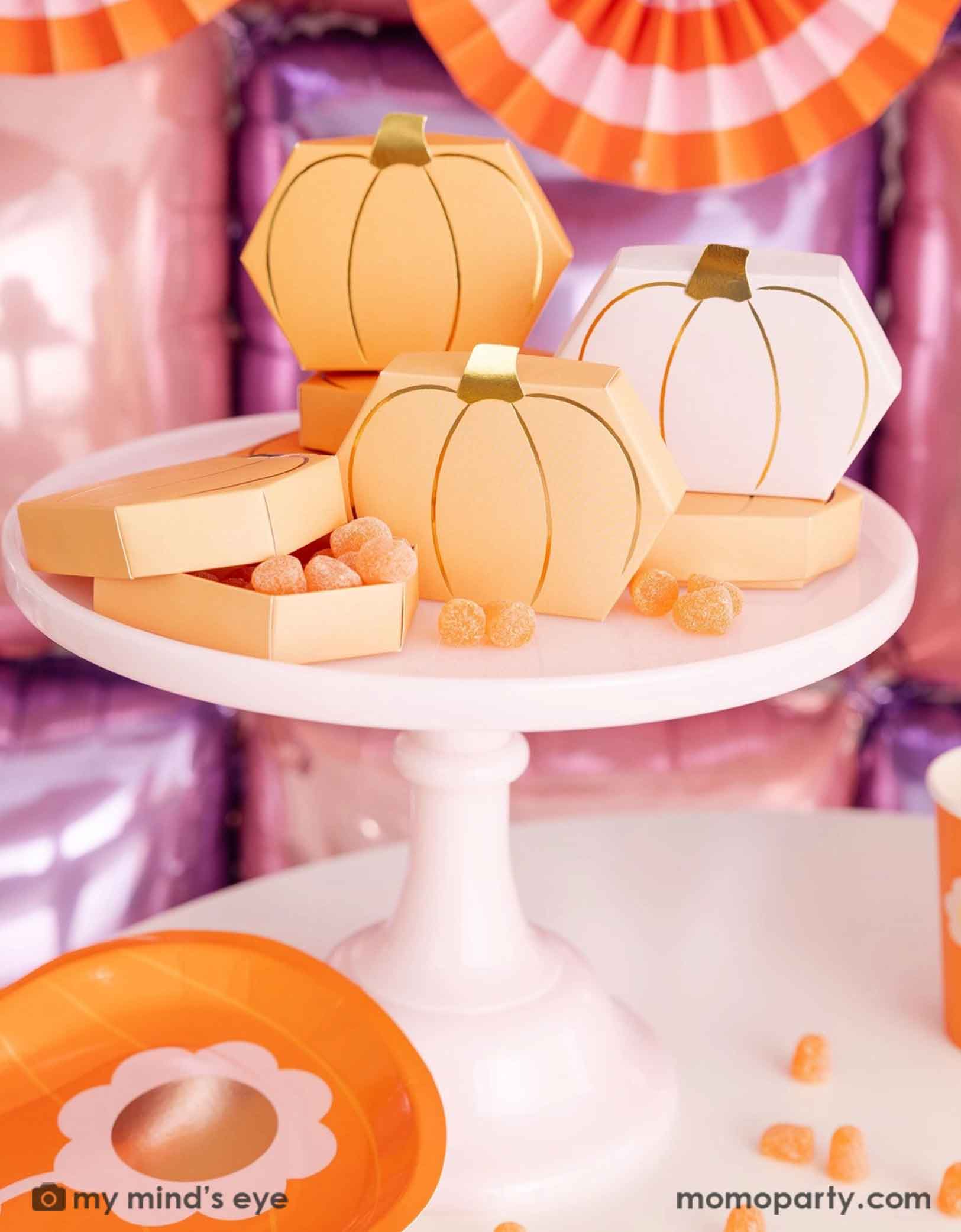 Momo Party's Pumpkin-shaped boxes by My Mind's Eye filled with orange gummies on a pale pink cake stand with festive pink Halloween decorative background with foil balloon wall and bunting garland.