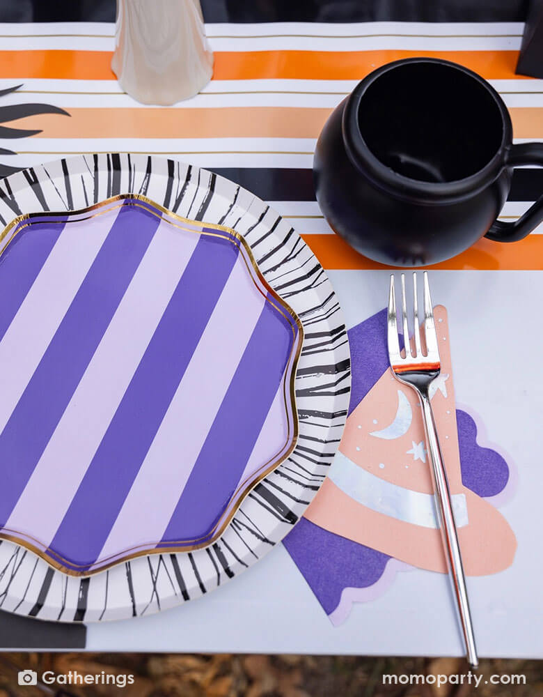 A close up shot of a festive Halloween witch themed party table featuring Momo Party's Bewitching purple side plates on a black and white striped dinner plates, bewitching witch shaped hat napkin and a cauldron shaped cup on a black and orange striped table runner. A perfect inspo for Halloween witch themed table for this spooky season!