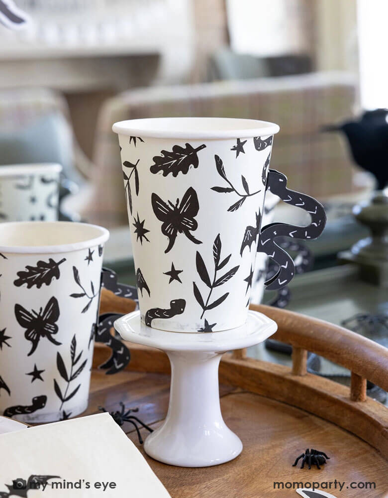 A close up shot of Momo Party's 12 oz Snake Handle Party Cups by My Mind's Eye on a wooden table covered with spider decoration and confetti. These spooky cups not only come in a fun snake handle design, but they will also make your party stand out in a unique way. Don't settle for boring party cups, add a touch of fun with these cups!