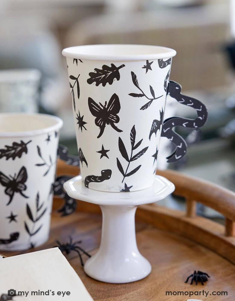 A close up shot of Momo Party's 12 oz Snake Handle Party Cups by My Mind's Eye on a wooden table covered with spider decoration and confetti. These spooky cups not only come in a fun snake handle design, but they will also make your party stand out in a unique way. Don't settle for boring party cups, add a touch of fun with these cups!