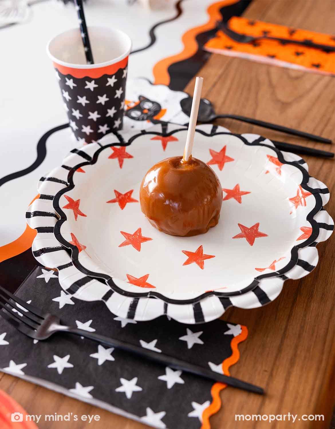 A Halloween-themed table setting featuring a caramel apple on a scalloped white paper plate with orange stars, alongside matching black and white star-patterned napkins, a star patterned black paper cup from Momo Party , and a fork on a wooden table.