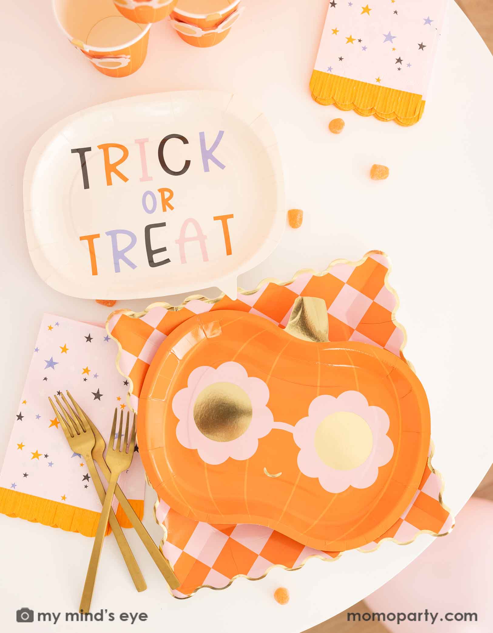 A Halloween-themed table featuring various pink Halloween themed tableware from Momo Party including a 'Trick or Treat' plate, a orange checkered dinner plate, a groovy pumpkin-shaped plate and party cups, plus pink star with orange fringed napkins. Making this an adorable tablescape for a cute Halloween party celebration.