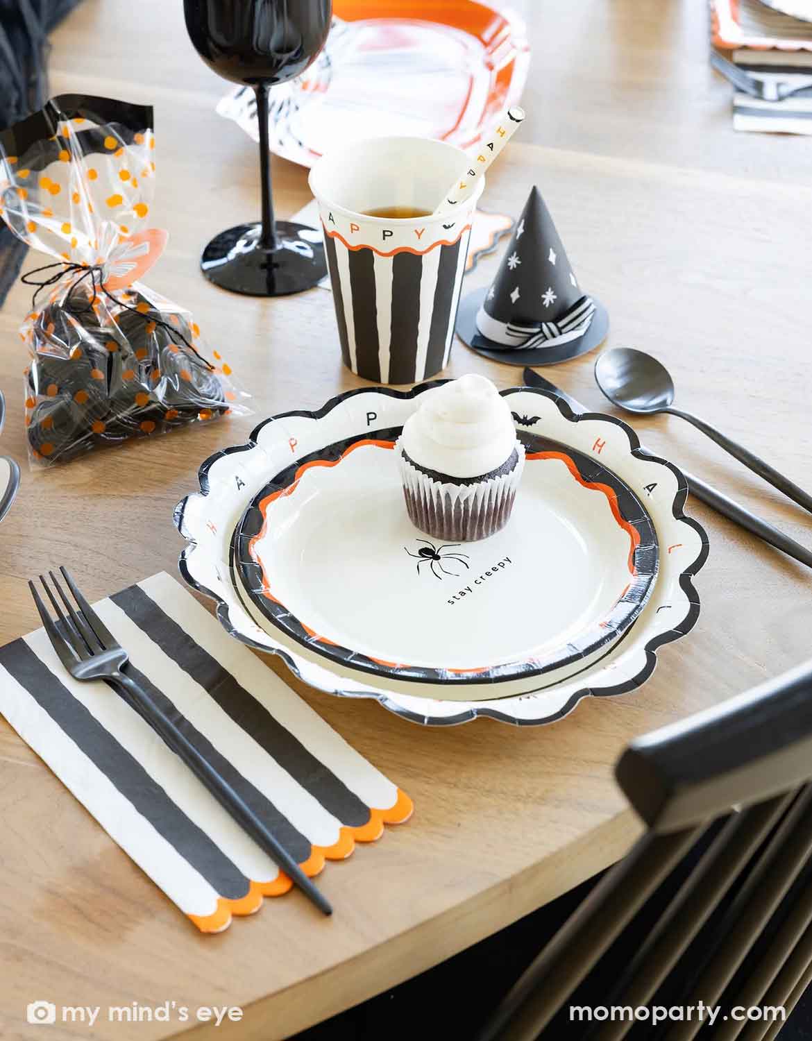 A styled Halloween table setting featuring multiple Halloween party supplies from Momo Party including a scalloped white plate with black and orange trim, a matching black and white striped napkin, a black witch hat shaped party hat treat holder, and a cup with black and white stripes, along with a treat bag with black candies on a wooden table.