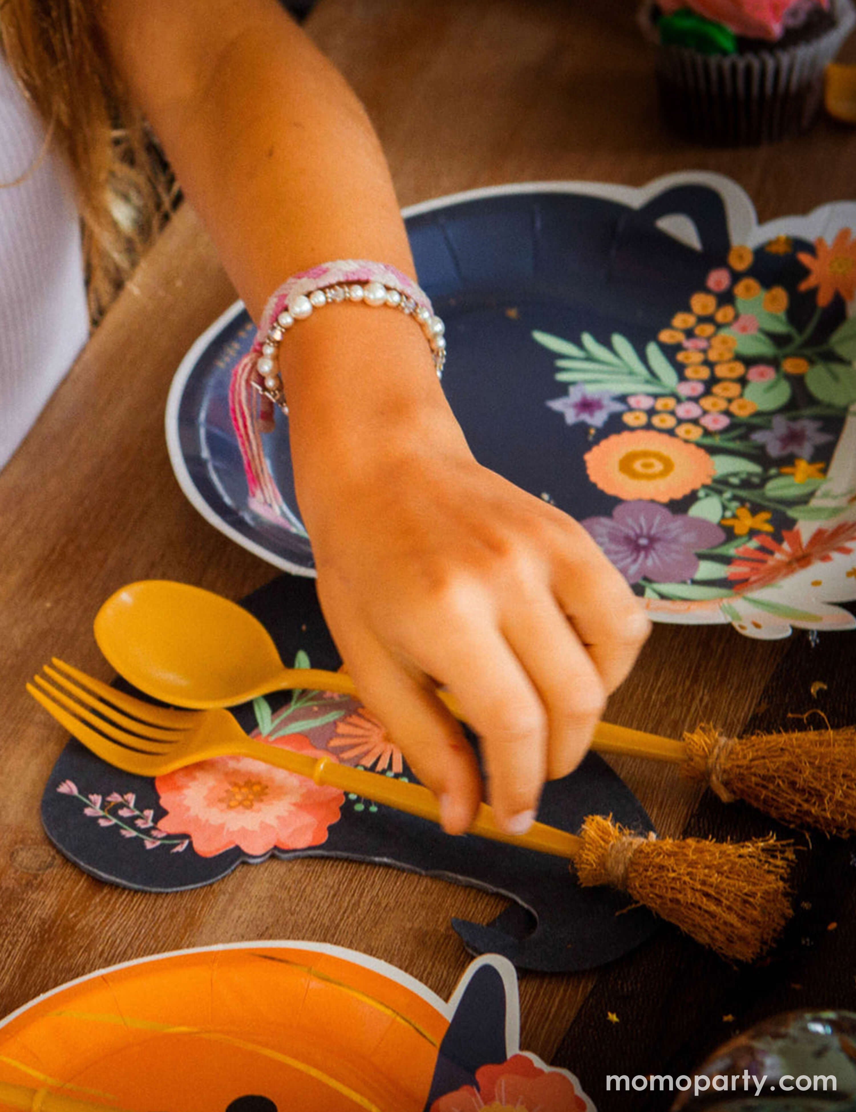 A girl's hand reaching Momo Party's witch's broom cutlery set on a witch themed Halloween party table featuring cauldron shaped party plates, pumpkin shaped party plates and witch hat shaped napkins with floral designs on them. Making this a great inspo for a boho and chic Halloween party tablescape.