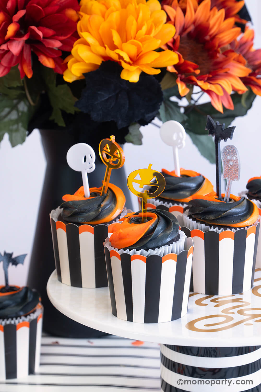 A close-up shot of some Halloween black and orange swirl cupcakes in Momo Party's Vintage Halloween striped food cups. The cupcakes are topped with glitter acrylic Halloween character shaped toppers including pumpkin, skull, ghost and bat. With some fall flower arrangement in the back, this gives a perfect inspo for a nostalgic Halloween celebration this spooky season!