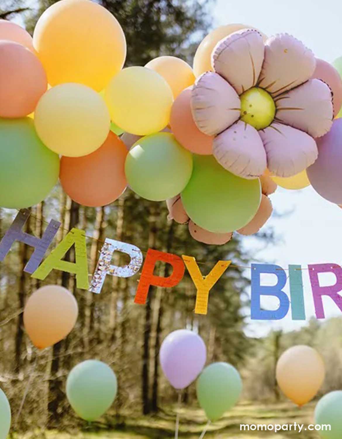 Colorful balloon arch in pastel colors with a lilac daisy-shaped foil balloon and Momo Party's 7ft 'Happy Birthday' banner in festive colors against a forest background.